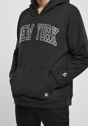 Osoba nosząca czarną bluzę z kapturem z napisem "NEW YORK" dużymi białymi literami na wysokości klatki piersiowej, z rękami w przedniej kieszeni.