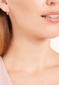 Ohrringe aus Roségold mit klaren, funkelnden Edelsteinen. Glatte Textur, runde Form und sichere Verschlussmechanik sichtbar gegen die Haut.