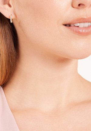 Boucles d'oreilles créoles en or rose avec des pierres précieuses transparentes et scintillantes. Texture lisse, forme ronde et fermeture sécurisée visibles sur la peau.