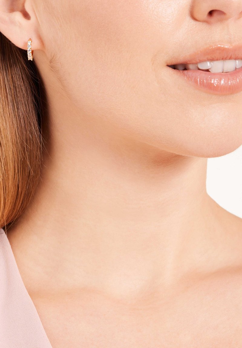 Ohrringe aus Roségold mit klaren, funkelnden Edelsteinen. Glatte Textur, runde Form und sichere Verschlussmechanik sichtbar gegen die Haut.