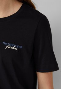 T-shirt in cotone nero con maniche corte, con la scritta "L'ESSENZA DELLA libertà" ricamata in blu e crema sul lato sinistro.