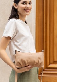 Beige leren clutch met een zachte, soepele textuur, ritssluiting en een afneembare band, vastgehouden door een persoon die een licht overhemd en groene broek draagt.