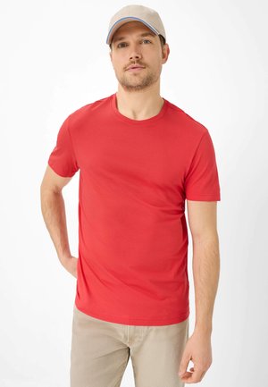 Uomo con cappellino beige, maglietta rossa a maniche corte e pantaloni beige, in piedi con una mano sui fianchi su sfondo bianco.