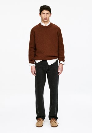 Młody mężczyzna stoi, ubrany w brązowy dzianinowy sweter na białą koszulę, czarne jeansy i beżowe sznurowane buty, na tle jednolitej białej ściany.