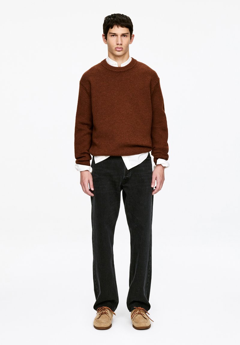Jeune homme debout portant un pull en maille marron par-dessus une chemise blanche, un jean noir et des chaussures à lacets couleur beige, sur un fond blanc uni.