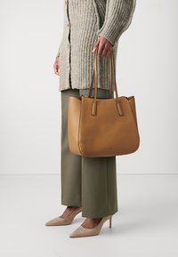 Sac fourre-tout en cuir marron avec une forme structurée, des poignées doubles et une texture subtile. Associé à un pantalon large vert olive et des talons beiges.