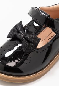 Czarne lakierowane buty z okrągłym noskiem, z perforowanymi detalami i teksturowaną czarną kokardką. Posiadają regulowany pasek i beżową podeszwę.