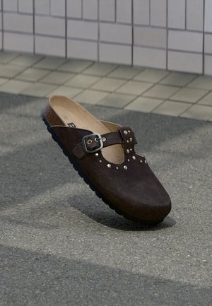 Zueco de ante marrón con punta redondeada, hebilla plateada y tachuelas decorativas plateadas a lo largo de la parte superior. Suela de goma negra.