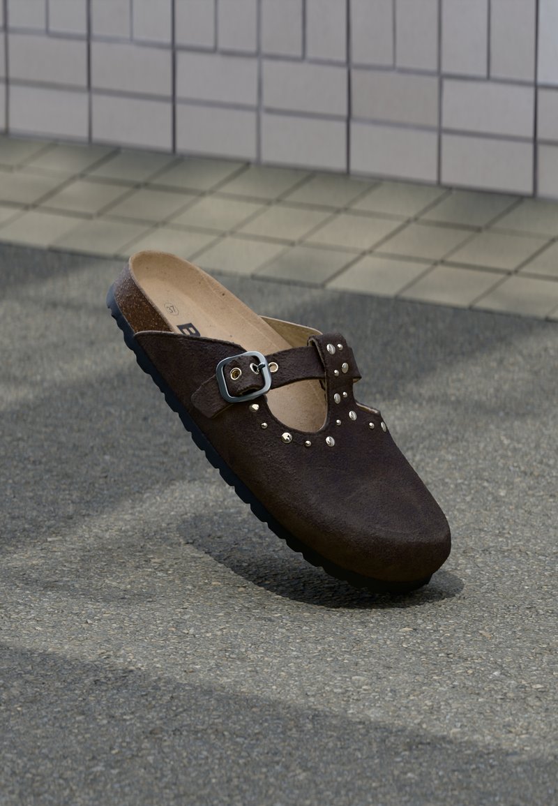 Καφέ σουέντ κλογκ με στρογγυλεμένη μύτη, ασημένια αγκράφα και διακοσμητικά ασημένια καρφιά στην επάνω πλευρά. Μαύρη ελαστική σόλα.