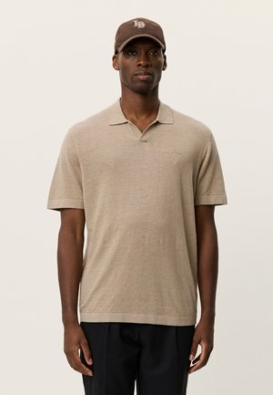 Polo beige a maniche corte con colletto, realizzata in un tessuto leggero, con un logo discreto del marchio sul petto. Indossata con un cappello marrone.