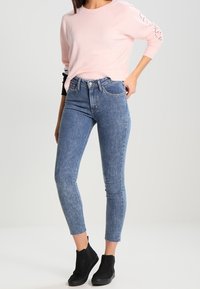 Kvinna som bär en ljusrosa långärmad topp, höga blå smala jeans och svarta ankelstövlar, stående mot en enkel ljus bakgrund.