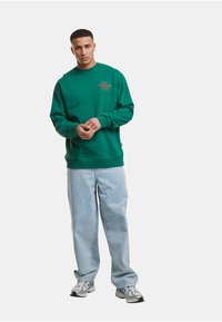 Sudadera verde con puños y dobladillo de canalé, que presenta un logo impreso en el pecho. Combinada con pantalones de mezclilla azul claro y zapatillas grises.