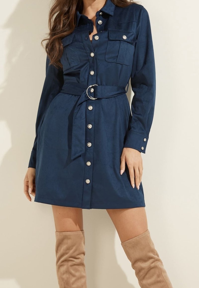 Femme portant une robe-chemise bleu marine ceinturée, à manches longues et deux poches poitrine, associée à des bottes en daim beige montant jusqu’au genou.