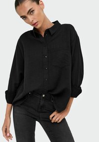 Camisa negra de botones en tejido ligero, con un bolsillo en el pecho, mangas remangadas y ajuste relajado, combinada con unos jeans negros.
