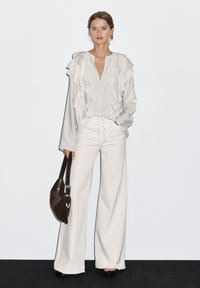 Woman wearing a white ruffled blouse and wide-leg pants, holding a dark brown leather handbag, standing against a white background.