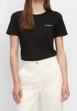 Frau trägt ein schwarzes T-Shirt mit kurzen Ärmeln und dem Schriftzug „(Re-Nature)“ auf der linken Brust sowie hoch taillierte, cremeweiße Hosen.