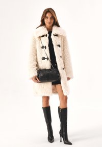 Cappotto in finta pelliccia crema con chiusure a toggles nere, abbinato a una borsa a tracolla nera e stivali neri con tacco a spillo al ginocchio.