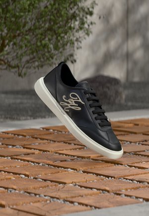 Pantof sport negru din piele, cu talpă albă și logo brodat bej pe lateral, plutind deasupra podelei din gresie de lemn, în aer liber, lângă verdeață.