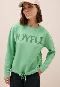 Sudadera verde con la palabra "JOYFUL" impresa en letras grandes. Presenta un escote redondo, un corte relajado y un cordón en el dobladillo.