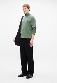 Light green zip-neck sweatshirt with embroidered logo, black bag, black trousers, and black shoes. Smooth fabric, relaxed fit, and long sleeves.