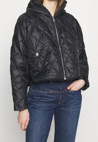 Nabíraná černá zkrácená bunda s kapucí, se zapínáním na přední zip a stříbrnými patentkami. Hladký povrch, lehký materiál.