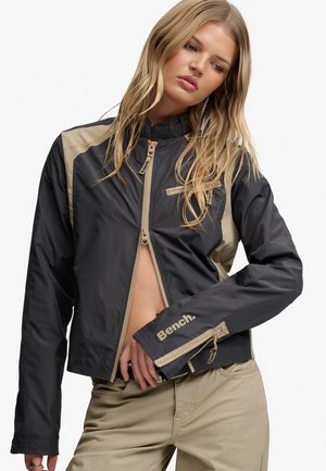 Junge Frau mit langen blonden Haaren, die eine schwarz-beige Bench-Jacke trägt, die bis zur Mitte des Oberkörpers geöffnet ist, und beige Hosen vor weißem Hintergrund.