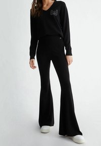 Pull noir à col en V avec détail de logo sur la poitrine gauche, associé à un pantalon noir évasé et des baskets blanches. Tissu doux en maille.