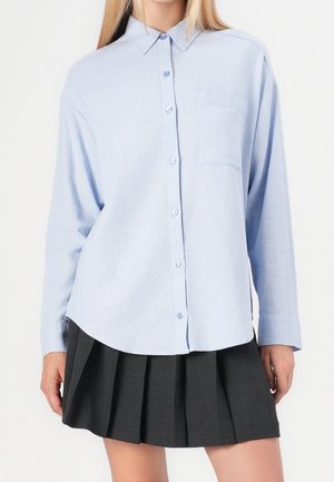 Frau trägt ein hellblaues Hemd mit Knöpfen und Brusttasche sowie einen dunkelgrauen, plissierten Rock vor einem einfarbigen Hintergrund.