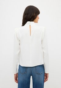 Blusa bianca a maniche lunghe con colletto alto, apertura a chiave sul retro e texture morbida. Abbinata a jeans di denim blu.