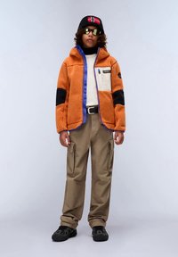 Asmuo vilkintis oranžine fliso striuke su kišenėmis, smėlio spalvos kargo kelnėmis, juodais batais, juoda kepure su raudona ir balta vėliava, ir atspindinčiais akiniais.