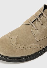 Chaussure beige en daim à lacets avec des perforations, un bout rond et une semelle en caoutchouc noire avec des accents de couture.