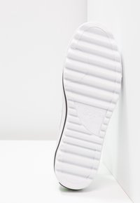 Podeszwa białego trampka Converse z wyraźnym wzorem ryflowanym i widocznym logo marki, ustawiona pionowo przy białej ścianie narożnej.