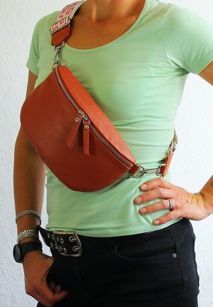 Braune Leder-Crossbody-Tasche mit texturierter Oberfläche, geschwungener Form, doppelten Reißverschluss-Anhängern und einem gemusterten verstellbaren Gurt. Um die Taille getragen.