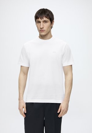 Uomo che indossa una t-shirt bianca semplice a maniche corte e pantaloni neri, in piedi davanti a uno sfondo bianco uniforme.