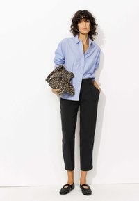 Chemise bleu clair à rayures, pantalon noir ajusté, écharpe à imprimé animal, et ballerines noires avec des embellissements. Cheveux bouclés, accessoires minimaux.