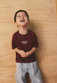 Burgundska bombažna majica z belim napisom »Neomejeni nasmehi vsak dan«, kombinirana s svetlo modrimi kavbojkami, stoji ob lesenem ozadju.