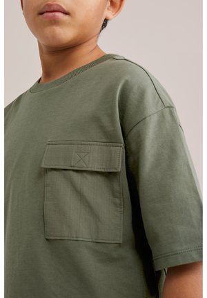 Zbliżenie osoby noszącej oliwkowozieloną koszulkę z dużą klapką kieszeni na lewej części klatki piersiowej.