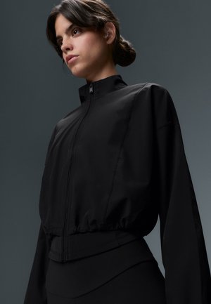Jeune femme aux cheveux foncés attachés, portant une veste noire zippée et un pantalon noir sur un fond gris.