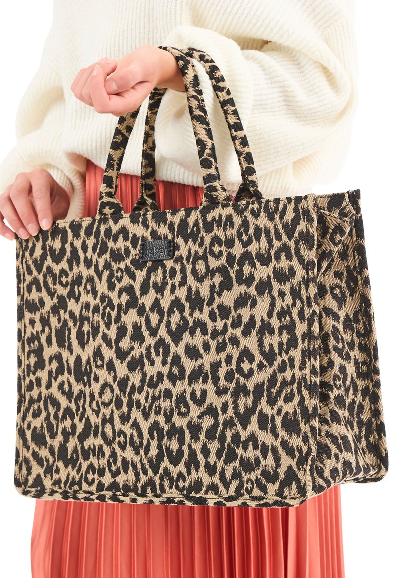 Personne tenant un grand sac fourre-tout à imprimé léopard avec des poignées doubles, portant un pull blanc et une jupe plissée rouge.