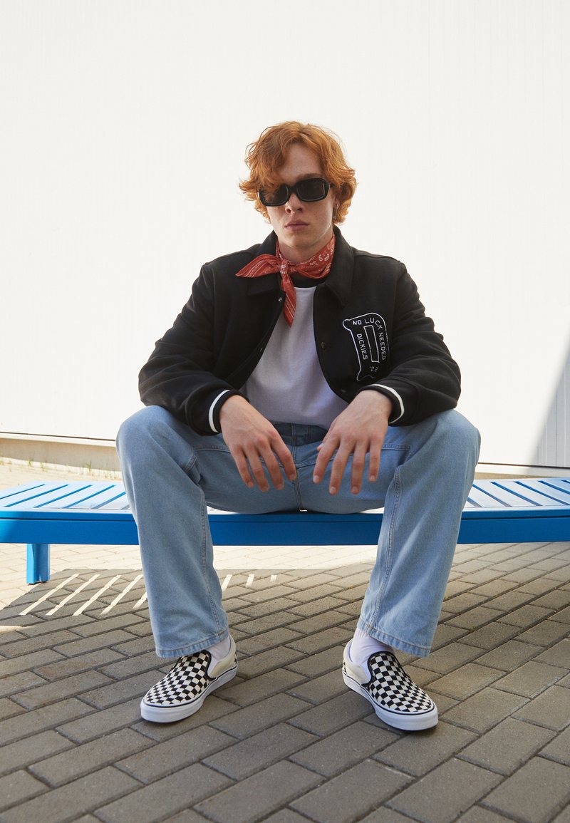 Black varsity jacket with white patch, white t-shirt, blue jeans, red bandana, and black-and-white checkered slip-on shoes, seated on blue bench.