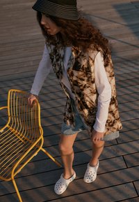 Chemise blanche à manches courtes sous un gilet oversize marron et crème à motifs, shorts en jean, sabots blancs et un chapeau seau rayé noir.