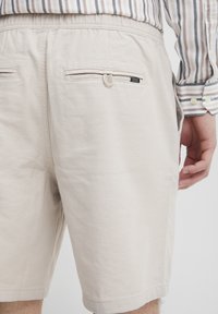Beige shorts laget av teksturert stoff, med elastisk midjebånd, to baklommer med knapper og et merke.