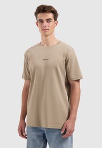 Jonge man in een beige "PURE PATH" t-shirt en lichtblauwe jeans, staand tegen een effen lichtgrijze achtergrond.