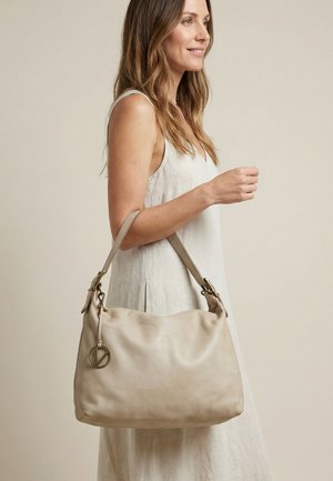 Žena v bezrukávových světle béžových šatech nesoucí velkou béžovou koženou kabelku přes rameno s kulatým kovovým přívěskem s logem.