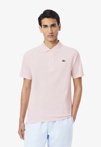 Jasnoróżowa koszulka polo z kołnierzykiem, trzema guzikami i zieloną haftowaną naszywką na piersi. Krótkie rękawy i gładka tekstura materiału.