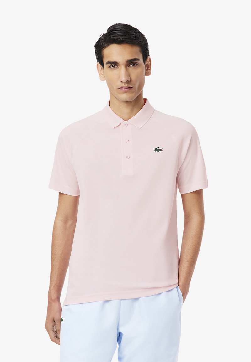 Jasnoróżowa koszulka polo z kołnierzykiem, trzema guzikami i zieloną haftowaną naszywką na piersi. Krótkie rękawy i gładka tekstura materiału.