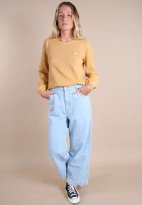 Un haut à manches longues texturé jaune moutarde associé à un jean large bleu clair. Des baskets noir et blanc viennent ajouter du contraste.