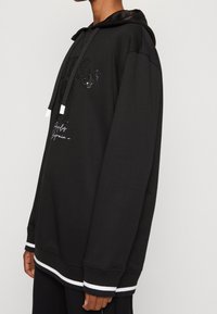 Sudadera con capucha negra con detalles de logotipo de lentejuelas, con cordones, puños de canalé y un dobladillo con rayas blancas. Hecha de material suave.