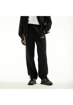 Persona che indossa pantaloni della tuta neri New Balance e sneakers abbinate, in piedi con una mano in tasca e una giacca sopra una canotta bianca.
