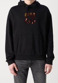 Sudadera negra con un bolsillo frontal, que presenta un logo reflectante "HU GO". El material es suave con una textura gruesa y tiene una capucha con cordón.